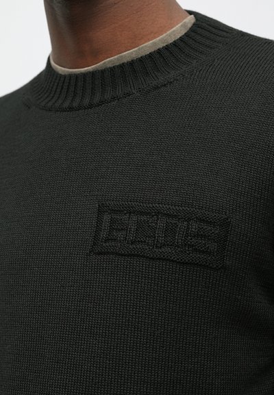 Artimas plano žmogaus, kuris dėvi juodą megztinį su šonkauliniais apykakle ir subtiliu pakeltu juodu "ELLES" logotipu ant krūtinės.