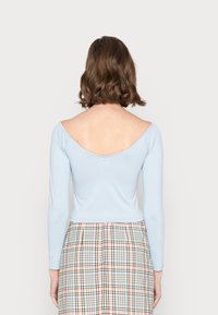 Camisola de manga longa azul claro com um design off-shoulder e costas arredondadas. Usada com uma saia xadrez em cores pastel e pregas estruturadas.