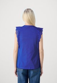 Žena s dlouhými blond vlasy má na sobě královsky modrý top bez rukávů s krajkovými volány, doplněný volnými modrými džíny, otočená zády.