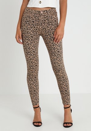 Femme portant un pantalon moulant motif léopard et des sandales noires à talons hauts, associées à un top blanc court.