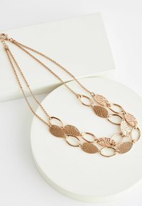 Collier en or rose avec des pièces en métal en forme de feuilles liées et ovales ; présente des motifs découpés et une fermeture à clip, reposant sur un présentoir blanc.