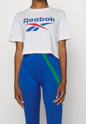 Osoba w białej krótkiej koszulce Reebok i niebieskich legginsach z wysokim stanem z zielonym ukośnym paskiem na lewym udzie.