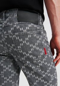 Vaqueros de mezclilla gris desgastados con un patrón de zigzag blanco repetitivo, etiqueta de cuero negra en la cintura y etiqueta de acento roja en el bolsillo trasero.