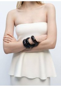Zwarte hars armbanden gestapeld op een arm, met een gladde textuur. Het model draagt een strapless witte top met een uitlopende zoom.