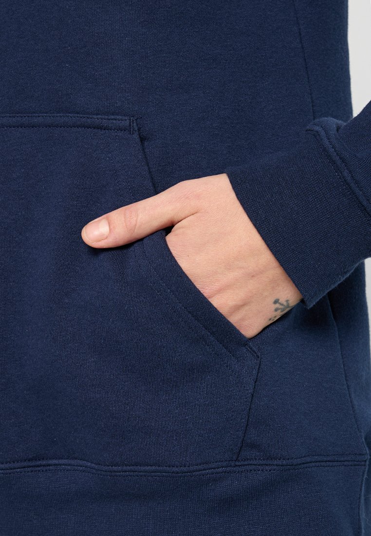 Mano con un piccolo tatuaggio sul polso all'interno della tasca anteriore di una felpa blu navy con polsini a coste.