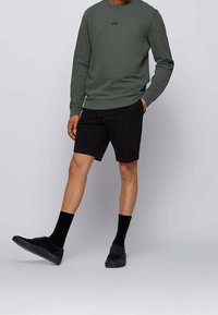 Sudadera verde oscura con cuello redondo, pantalones cortos negros, calcetines negros y zapatillas negras. Diseño simple con un mínimo de marca.