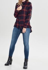 Red and navy plaid button-up shirt with a collar, long sleeves featuring embellishments, paired with blue skinny jeans and black ankle boots.