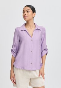 Camicia button-up di un leggero lavanda, con colletto, maniche tre quarti e una vestibilità morbida. Presenta un tessuto testurizzato e due tasche frontali.