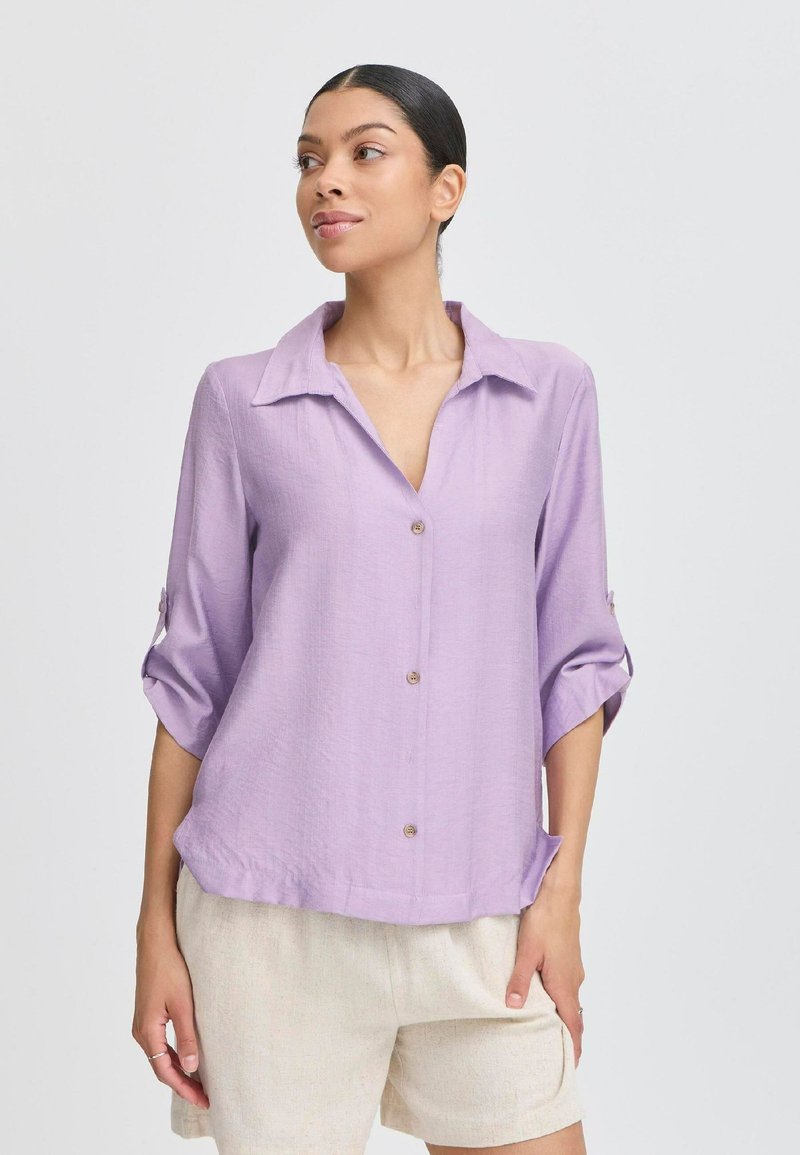 Camicia button-up di un leggero lavanda, con colletto, maniche tre quarti e una vestibilità morbida. Presenta un tessuto testurizzato e due tasche frontali.