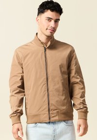 Chaqueta bomber marrón con cierre de cremallera, cuello alto y puños elásticos. Presenta una textura suave y un diseño minimalista.