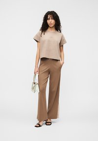 Μπεζ κοντομάνικη μπλούζα με διακριτικό τύπωμα, σε συνδυασμό με καφέ παντελόνια φαρδιάς γραμμής με σκισίματα, και μια ανοιχτοπράσινη τσάντα. Μαύρα σανδάλια.