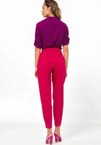 Dopasowana fioletowa koszula z podwiniętymi rękawami połączona z różowymi spodniami o wysokim stanem. Różowe strappy obcasy dopełniają outfit. Gładkie białe tło.