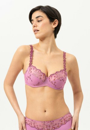 Femme portant un soutien-gorge et un slip rose assortis brodés de fleurs, regardant sur le côté sur un fond clair uni.