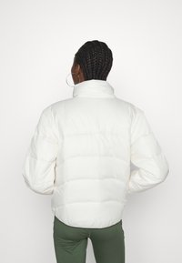 Biała puchowa kurtka z wysokim kołnierzem, z poziomymi przeszyciami i logo z tyłu. Teksturowany materiał, lekka konstrukcja.