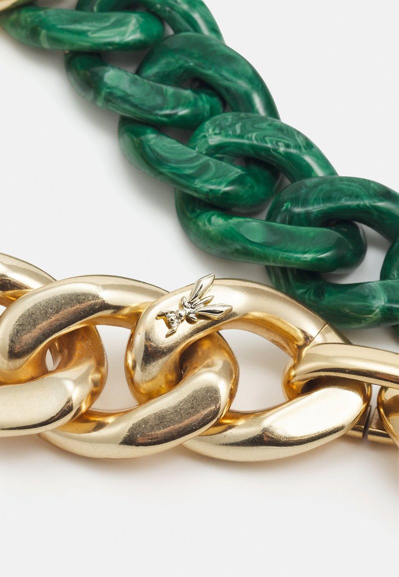 Eine massige goldene Gliederkette mit einem kleinen Bienenmotiv liegt unter einer massiven marmorierten grünen Gliederkette auf einem weißen Hintergrund.