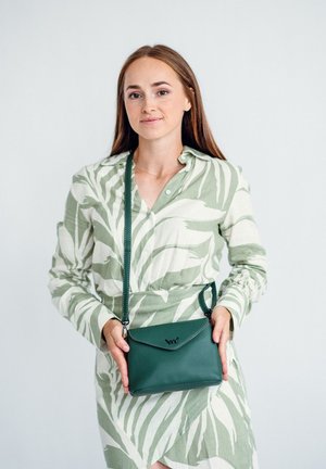 Grüne Leder-Crossbody-Tasche mit einer umklappbaren Klappe und geprägtem Logo. Getragen mit einem hellgrünen und weißen gemusterten Hemd.