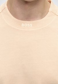 Gros plan d'un t-shirt beige clair à col rond avec le texte brodé en relief "BOSS" au centre de l'encolure avant, sur la partie supérieure de la poitrine d'une personne.