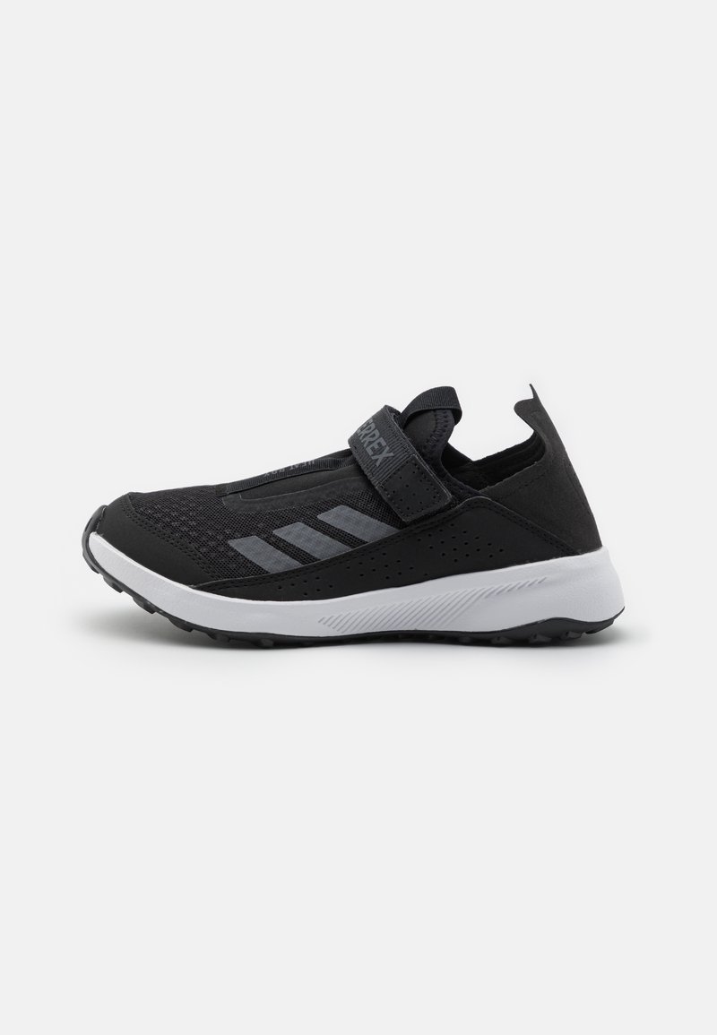 Schwarzer Sportschuh mit einem Mesh-Obermaterial, Klettverschluss, grauen Akzenten und einer weißen Sohle. Merkmale sind strukturierte Details und ein niedriges Profil.