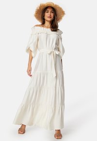 Hvid maxi kjole med off-the-shoulder halsudskæring, bølget kant, korte puffærmer, bundet talje og lagdelt nederdel. Sammen med en stråhat.