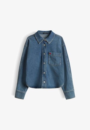 Giacca di denim blu con maniche lunghe, bottoni frontali, taschino sul petto e un piccolo dettaglio a cuore rosso sulla tasca.