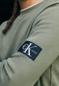 Olivgrön långärmad skjorta i texturerad tyg med en svart patch med "Calvin Klein Jeans"-logotyp på vänster ärm.