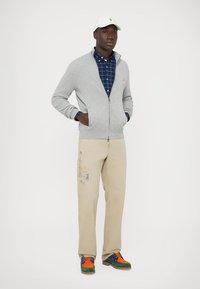 Cardigã cinza claro, camisa azul aos quadrados, calças bege com estampa gráfica e sapatos multicoloridos (laranja, verde, azul) usados por uma figura masculina.