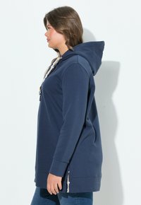 Námořnická modrá mikina s klokánkovou kapsou, kapucí se stahovací šňůrkou a postranními rozparky. Vyrobena z měkkého materiálu s volným střihem a dlouhými rukávy.