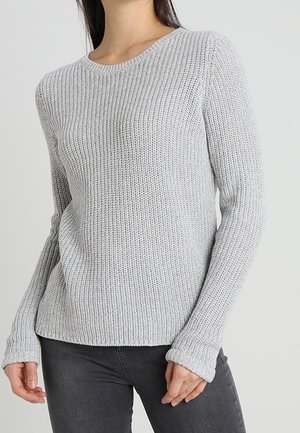 Femme portant un pull en maille côtelée gris clair à manches longues et un pantalon ajusté gris foncé, debout devant un fond uni.