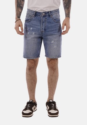 Hombre que lleva pantalones cortos de mezclilla azul deslavada con detalles desgastados y zapatillas en blanco y negro, de pie con los brazos relajados a los lados.