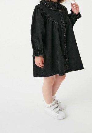 Robe en jean noire avec un col à volants, des manches longues avec des poignets élastiques, et un devant boutonné. Associée à des baskets blanches à scratch.