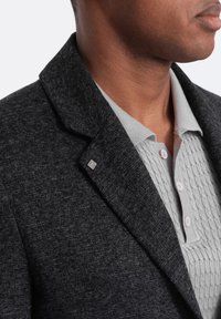 Blazer texturé gris foncé avec un col cranté, arborant un petit logo argenté. En dessous, un polo gris clair à motifs avec des boutons.