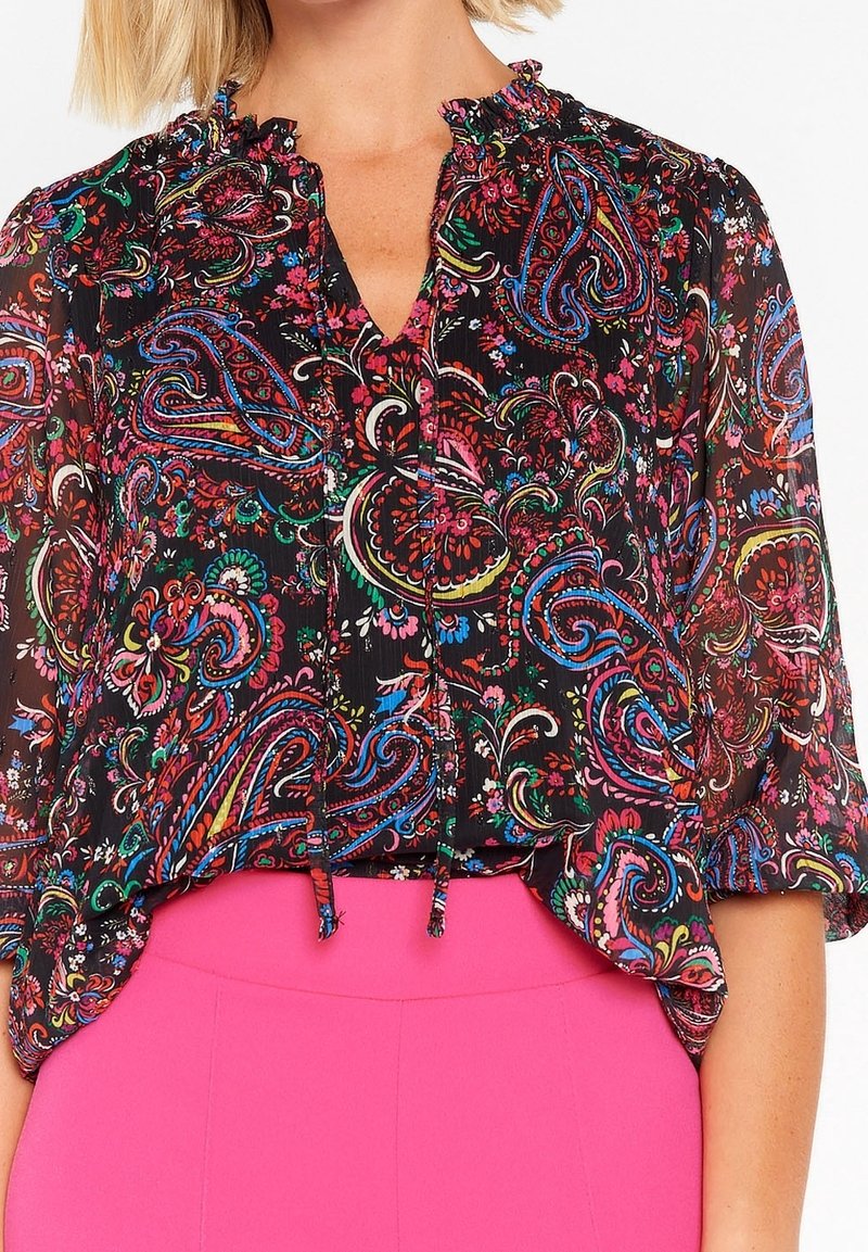 Femme portant un chemisier noir transparent avec des motifs paisley colorés et floraux, associé à un pantalon taille haute rose vif.