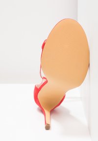 Chaussure à talon haut en suède rouge vue de dos, avec un talon aiguille et une semelle beige lisse ornée d'un motif texturé.