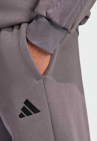 Šedé sportovní tepláky s měkkou texturou, které mají boční kapsu a černé logo Adidas blízko spodní části. Hribovaný lem u kotníku.