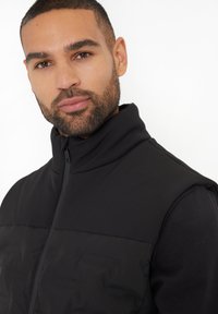 Schwarze ärmellose Steppweste mit hohem Kragen, aus glattem Material und mit Reißverschluss, über einem schwarzen Langarmshirt getragen.
