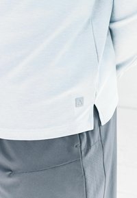 Gaiši zila krekla apakšmala ar mazu logotipu virs šķēlēm, savienota ar pelēkām biksēm, kas izceļas ar izšūtu kabatu un perforētu sānu paneli.