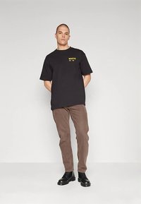 Crna majica s žutim logotipom "MAKIA", uparena s smeđim hlačama i crnim cipelama sa vezicama. Jednostavan dizajn, opušten kroj i mekana tekstura.