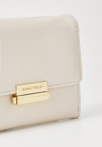 Kremfarget teksturert lommebok med et clasp i gulltone. Har en debossed "ANNA FIELD" logo på forsiden. Kompakt rektangulær form.