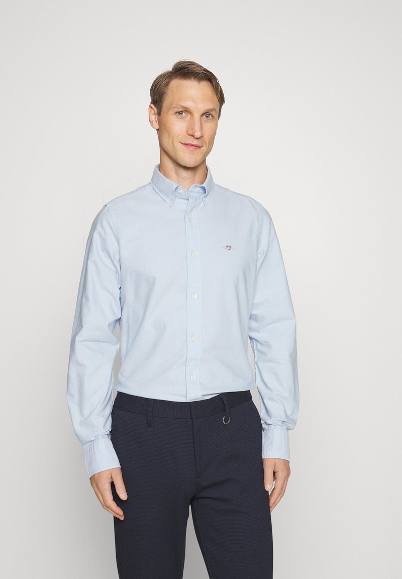 Homme portant une chemise bleu clair à boutons et un pantalon foncé, debout devant un fond clair uni.