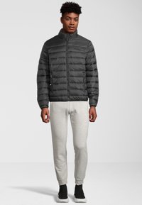 Vadderad svart jacka med hög krage, horisontell quiltning och framtida dragkedja. Bärs över ljusgrå sweatpants och svarta skor.