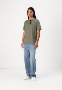 T-shirt verde oliva con maniche corte, scollatura rotonda e dettaglio del logo; abbinato a jeans baggy azzurri e sneakers bianche.