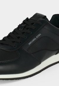 Baskets noires Michael Kors avec panneaux en cuir et tissu, lacets noirs et semelle blanche, avec détail du logo sur le côté.