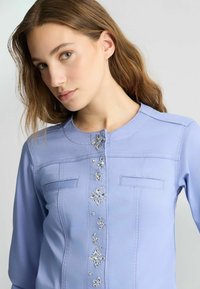 Helles blauer Bluse mit einem runden Halsausschnitt, zwei Brusttaschen und dekorativen Strassverzierungen auf der Vorderseite. Glatte Stoffstruktur.