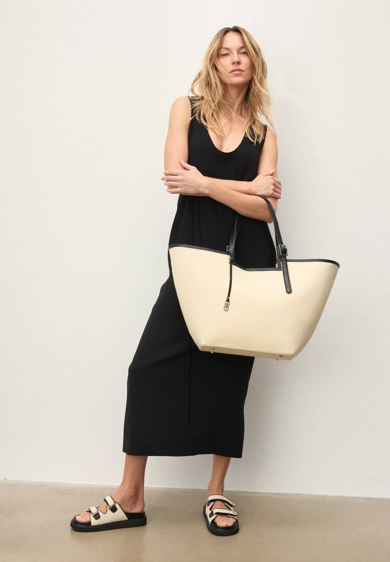 Grande borsa tote beige con accenti neri, forma elegante, manici robusti. Modella che indossa un abito nero e sandali beige e neri.