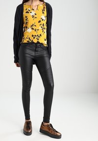 Pantalons en cuir synthétique noir avec des détails en relief, associés à un haut floral jaune et un cardigan sombre. Des baskets marron complètent la tenue.