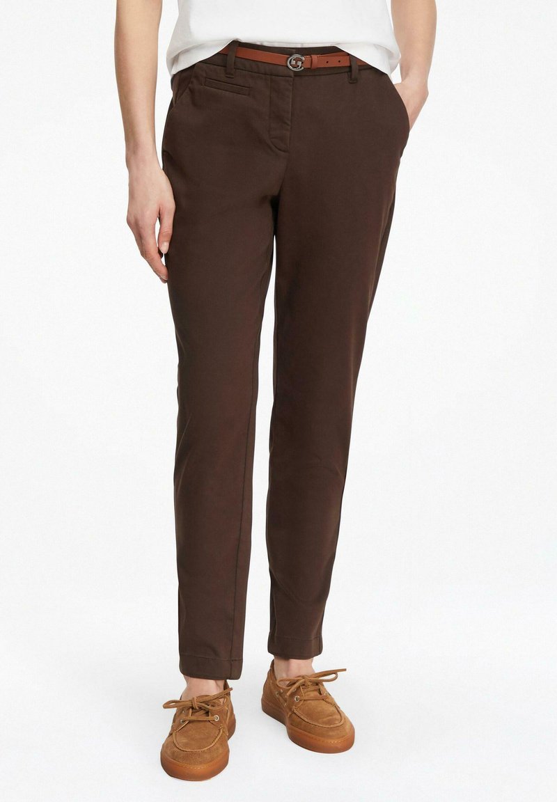 Osoba ubrana w ciemnobrązowe spodnie o dopasowanym kroju, jasnobrązowy skórzany pasek i brązowe zamszowe buty sznurowane na tle białego tła.