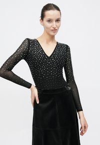 Μαύρο κορμάκι μακρυμάνικο με V-λαιμόκοψη, διακοσμημένο με ασημί καρφιά. Συνδυασμένο με μαύρη υφασμάτινη φούστα με λεία επιφάνεια.