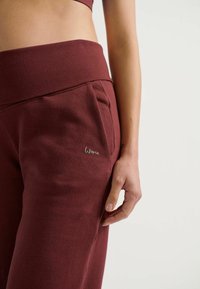 Bordeaux sweatpants met een brede tailleband, zijzakken en een subtiele 'Woma' logo in zilveren stikken. Zacht, getextureerd materiaal.