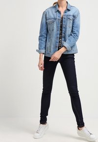 Chaqueta de denim azul claro con dos bolsillos en el pecho, cierre de botones y mangas remangadas, combinada con jeans skinny oscuros y zapatillas blancas.