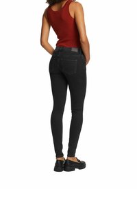 Femme portant un jean skinny noir, un haut rouge sans manches côtelé et des mocassins noirs, debout, les mains dans les poches arrière, dos tourné.